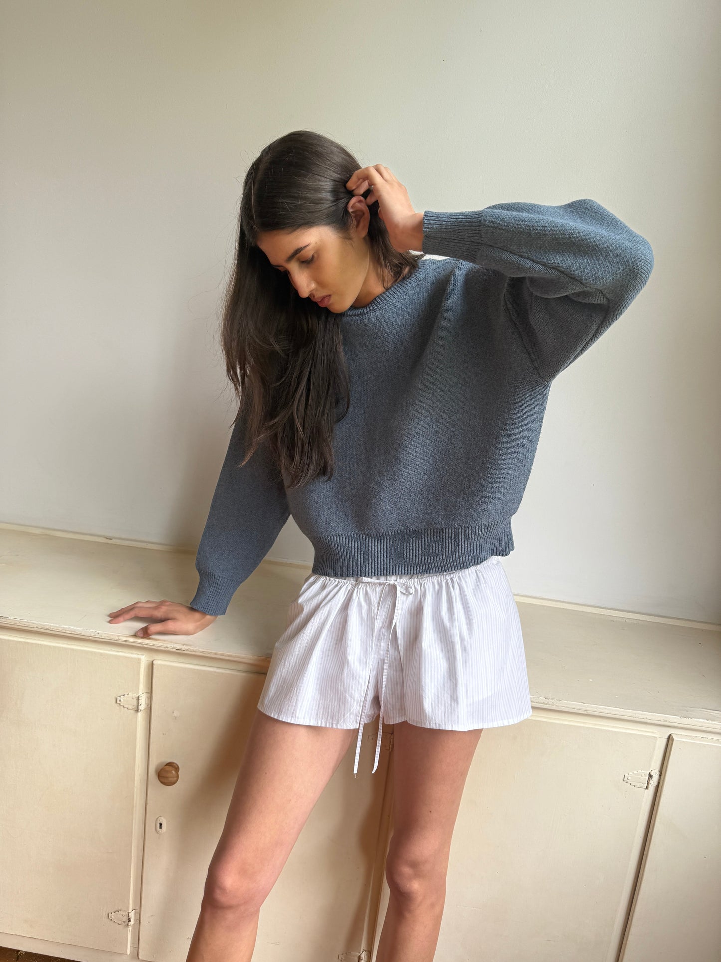A woman with long dark hair wears the 0 Pullover in Glacier Blue with white drawstring shorts. She stands indoors, looking down, one hand touching her head and the other resting on a light-colored cabinet.