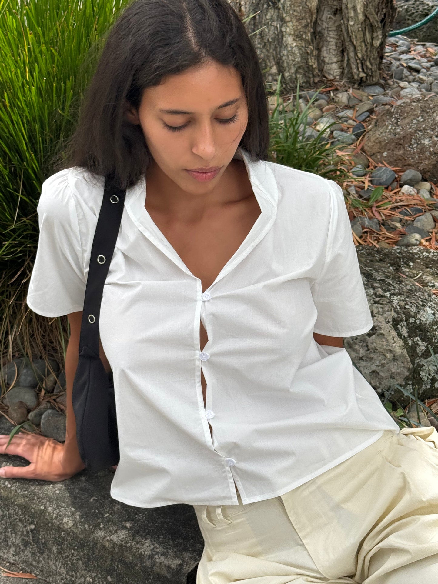 A person with long dark hair sits outdoors on a rock, wearing the Sails Top - White from Deiji Studios and light-colored pants, looking down with a black bag over one shoulder amid lush greenery and rocks.