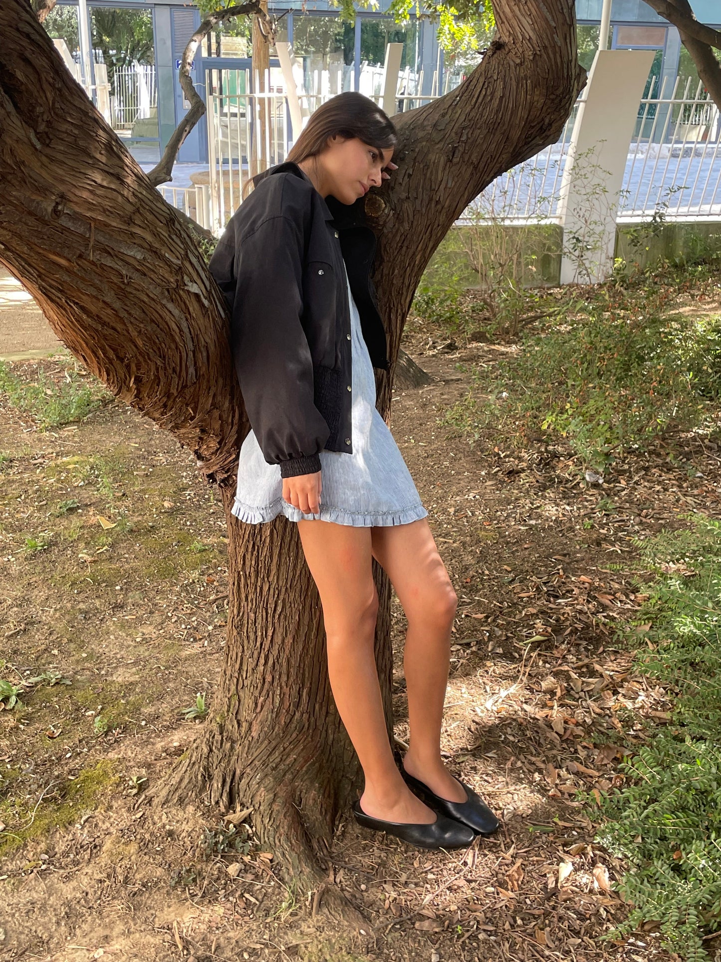 Wearing the Shirring Jacket - Slate over a light denim dress and black flats, a person leans against a garden tree, eyes closed and one leg bent on the trunk, as sunlight filters through the leaves above.