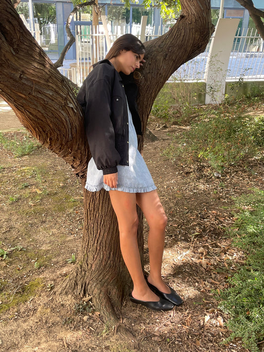 Wearing the Shirring Jacket - Slate over a light denim dress and black flats, a person leans against a garden tree, eyes closed and one leg bent on the trunk, as sunlight filters through the leaves above.
