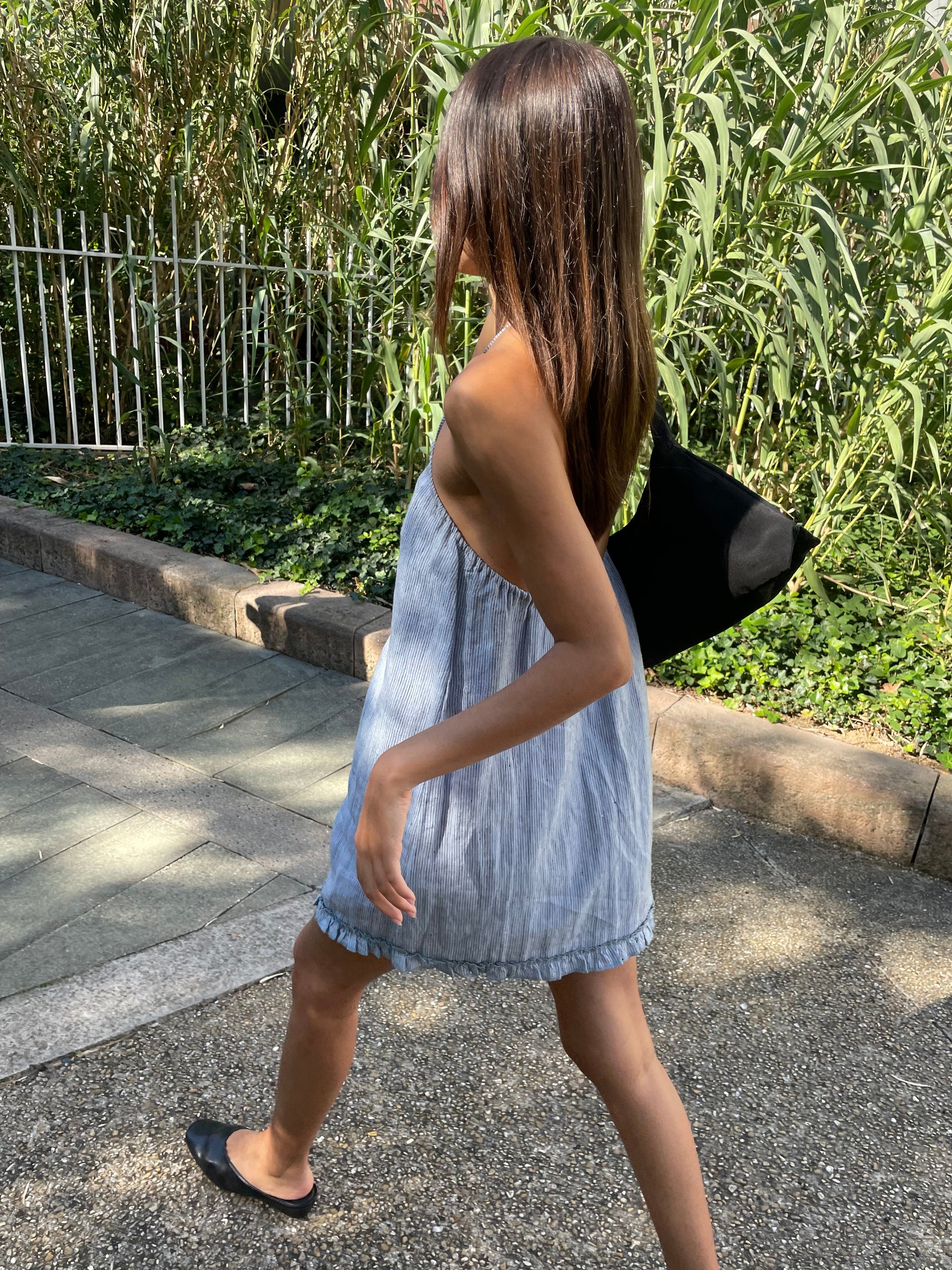 A woman with long brown hair walks outside along a path, wearing a light blue Flounce Halter Dress - Blue Stripe by Deiji Studios, black bag, and black flat shoes. Tall green plants and a white fence line the background.