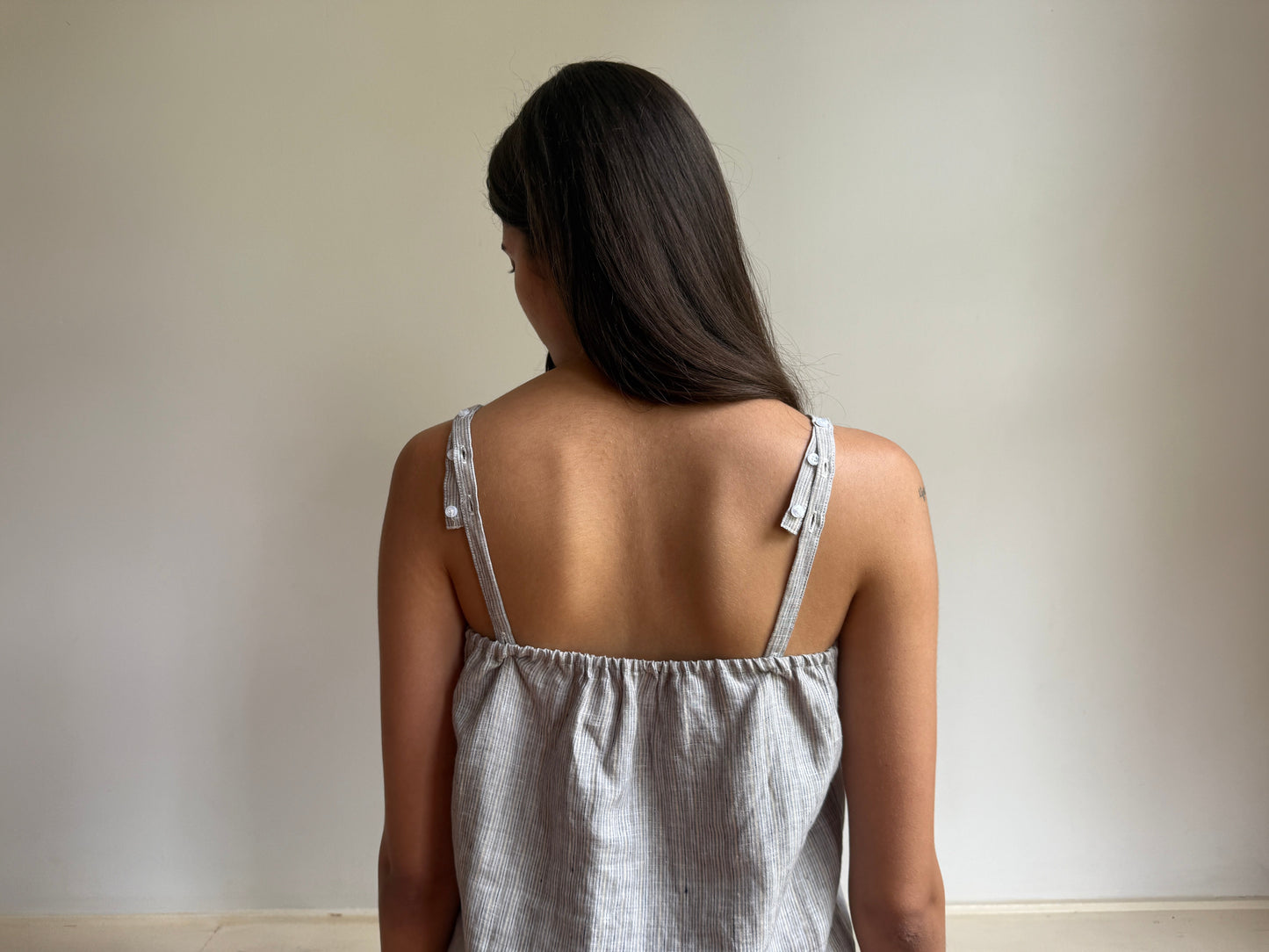 A woman with long dark hair is seen from behind, wearing the 0 Button Linen Set in Haze Blue, featuring a sleeveless design with thin straps, as she stands against a plain light-colored wall.