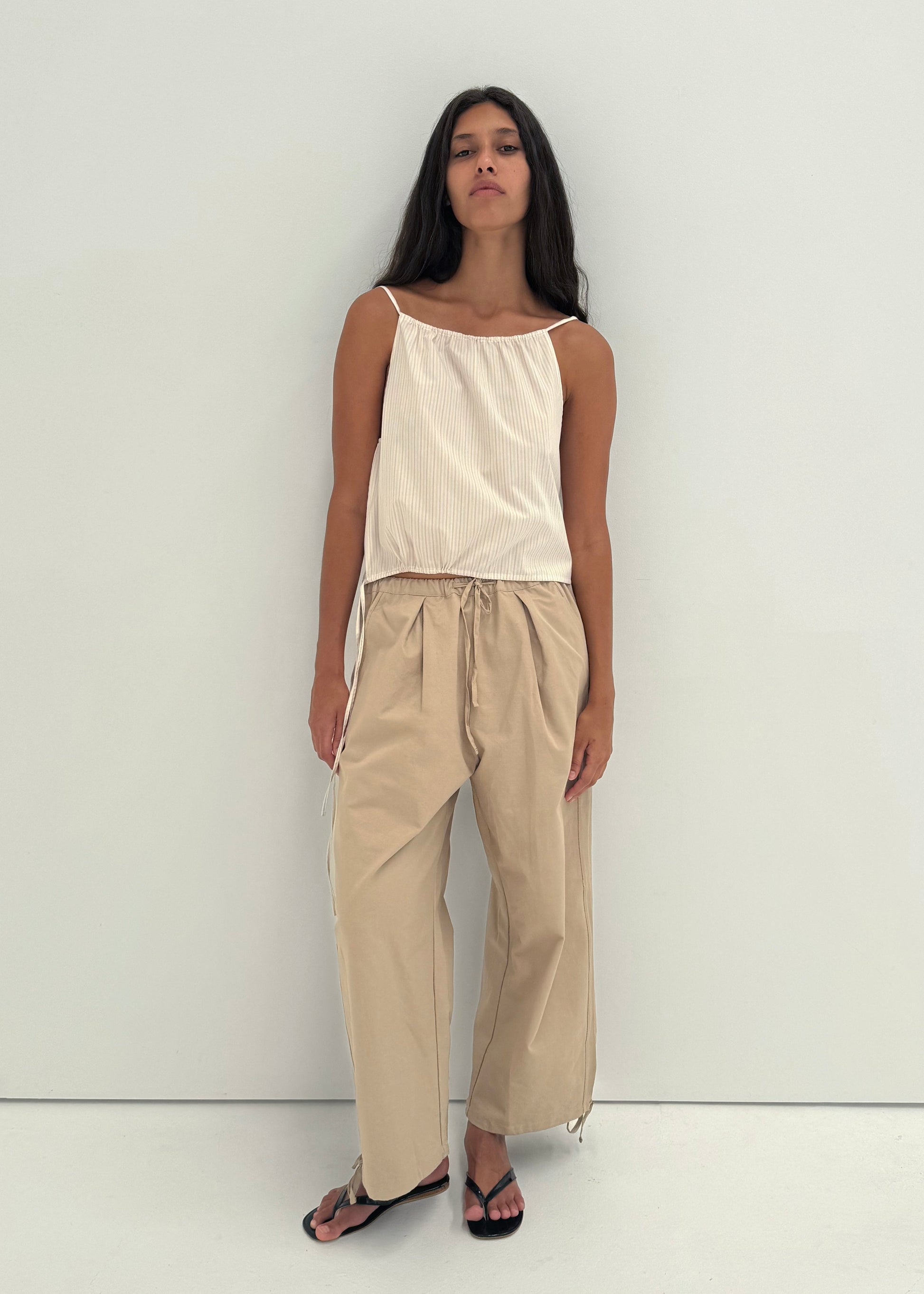 A woman with long dark hair stands against a white wall, wearing the Drawcord Wide Leg Pant - Cream in organic cotton with a beige sleeveless top and black sandals. She has a neutral expression, arms at her sides.