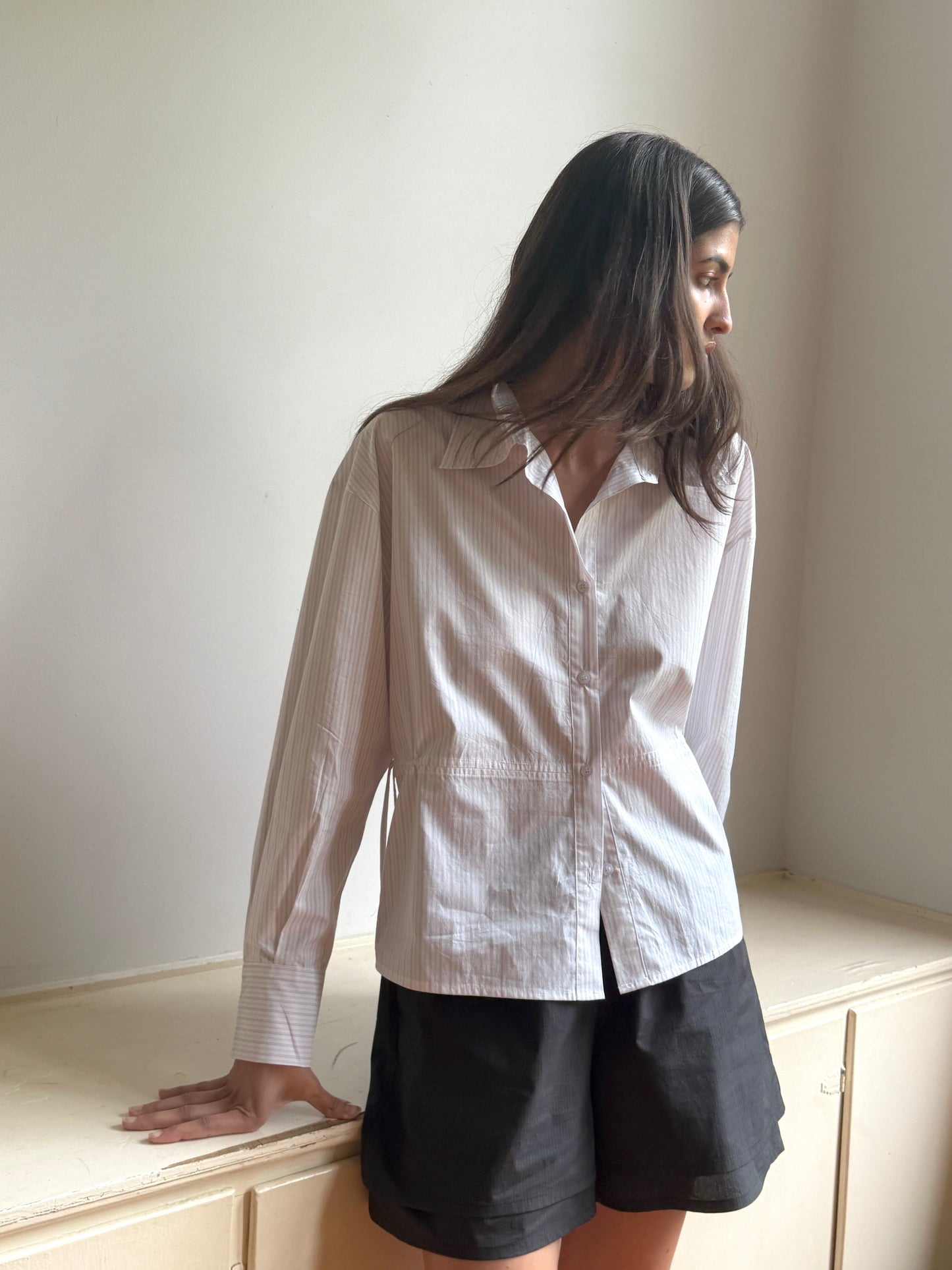 A woman with long dark hair wears the 0 Layered Short - Black Stripe and a light striped button-up shirt, leaning against a light cabinet and looking to the side in gentle lighting.