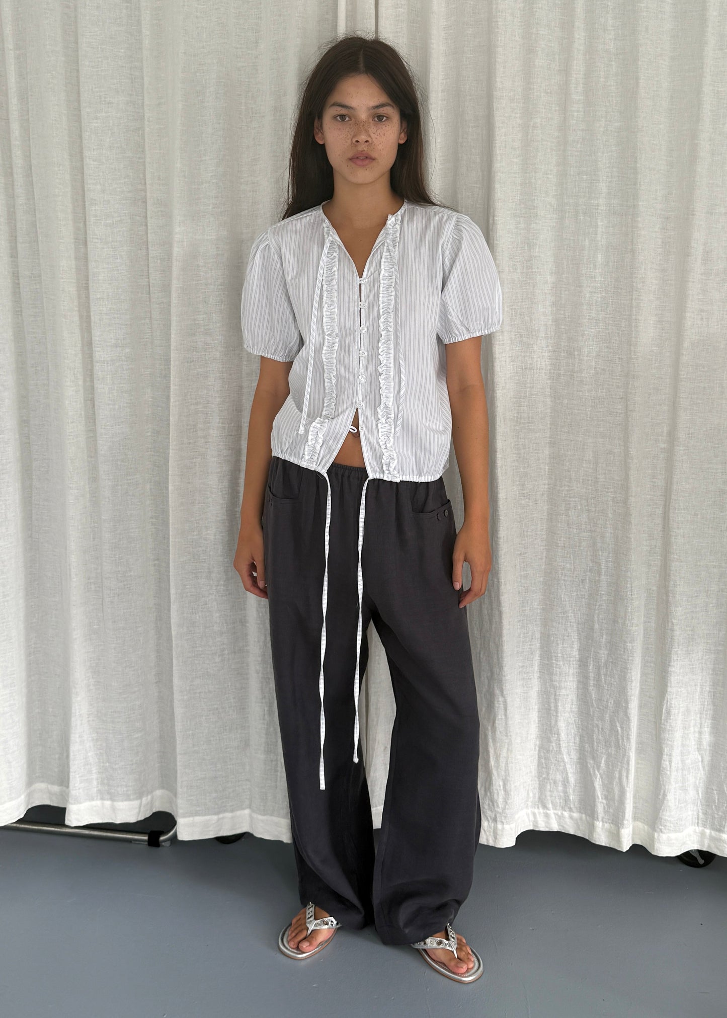 A woman stands before a white curtain, wearing the Double Frill Top - Sage Stripe by Deiji Studios with black wide-leg drawstring pants and sandals. She gazes at the camera with a neutral expression.