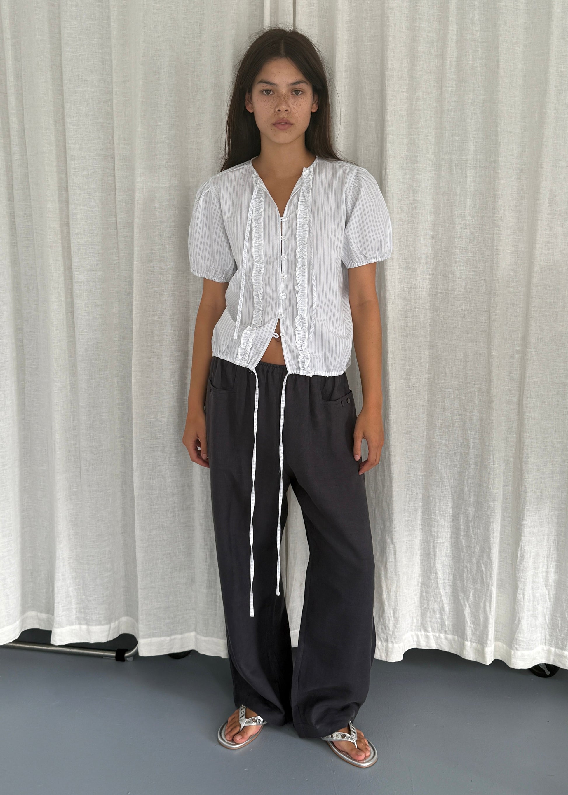 A woman stands before a white curtain, wearing the Double Frill Top - Sage Stripe by Deiji Studios with black wide-leg drawstring pants and sandals. She gazes at the camera with a neutral expression.