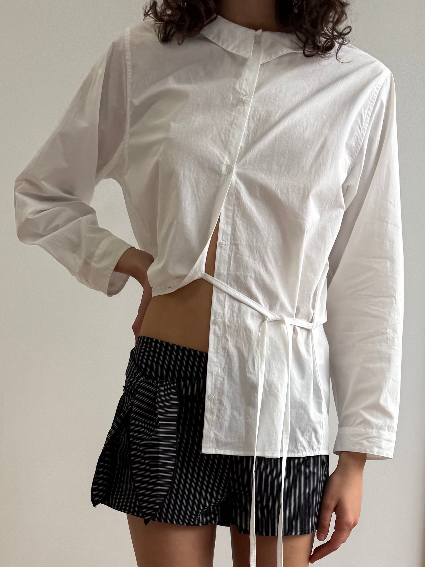 A person wearing the Left Shirt, a white asymmetrical wrap blouse with long sleeves, paired with black pinstriped shorts. The blouse is partially open, showing a hint of midriff. The person's head is not fully visible.