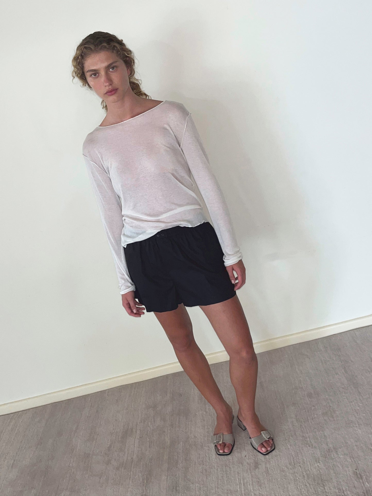 A person with curly hair stands indoors against a white wall, wearing a sheer white long-sleeve shirt, Organic Cotton Boxer - Black shorts, and gray slide sandals. They have a relaxed posture and a neutral expression.