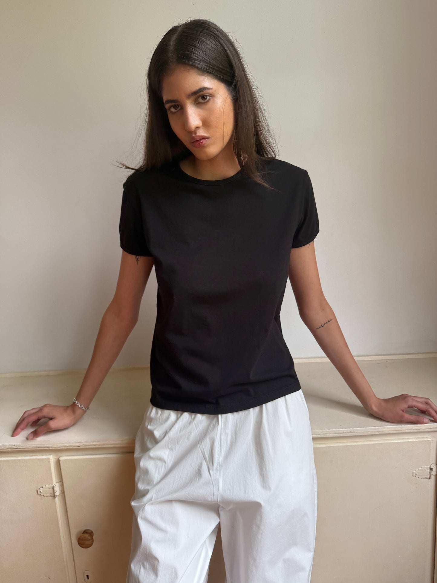 A woman with straight dark hair models the 0 Simple Tee - Black, paired with relaxed fit white pants, standing against a light wall, hands on a cabinet, and looking at the camera with a neutral expression.