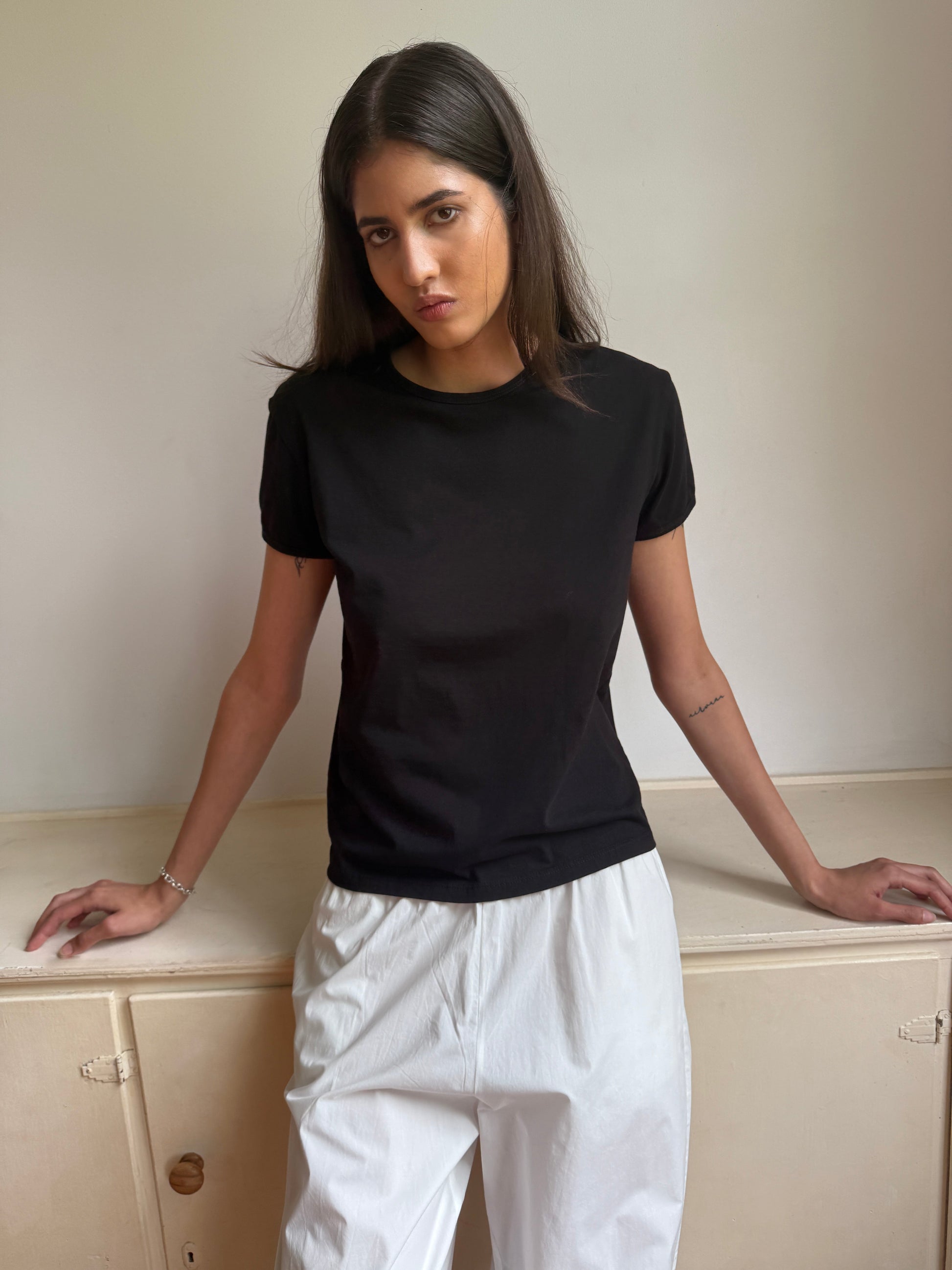 A woman with straight dark hair models the 0 Simple Tee - Black, paired with relaxed fit white pants, standing against a light wall, hands on a cabinet, and looking at the camera with a neutral expression.