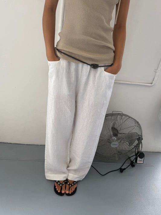 A person stands indoors with their hands in the pockets of Linen Square Pocket Pants, styled with a beige sleeveless top and black sandals. A large floor fan is nearby against the wall.