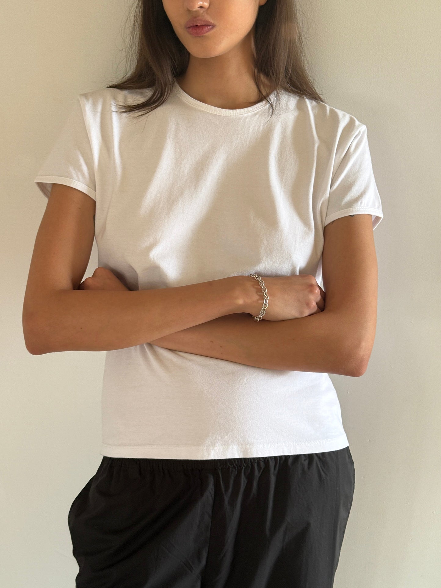 Model wears the 0 Simple Tee - White, paired with black pants, standing with arms crossed in front of a light wall. Only their upper body and part of their face are visible.