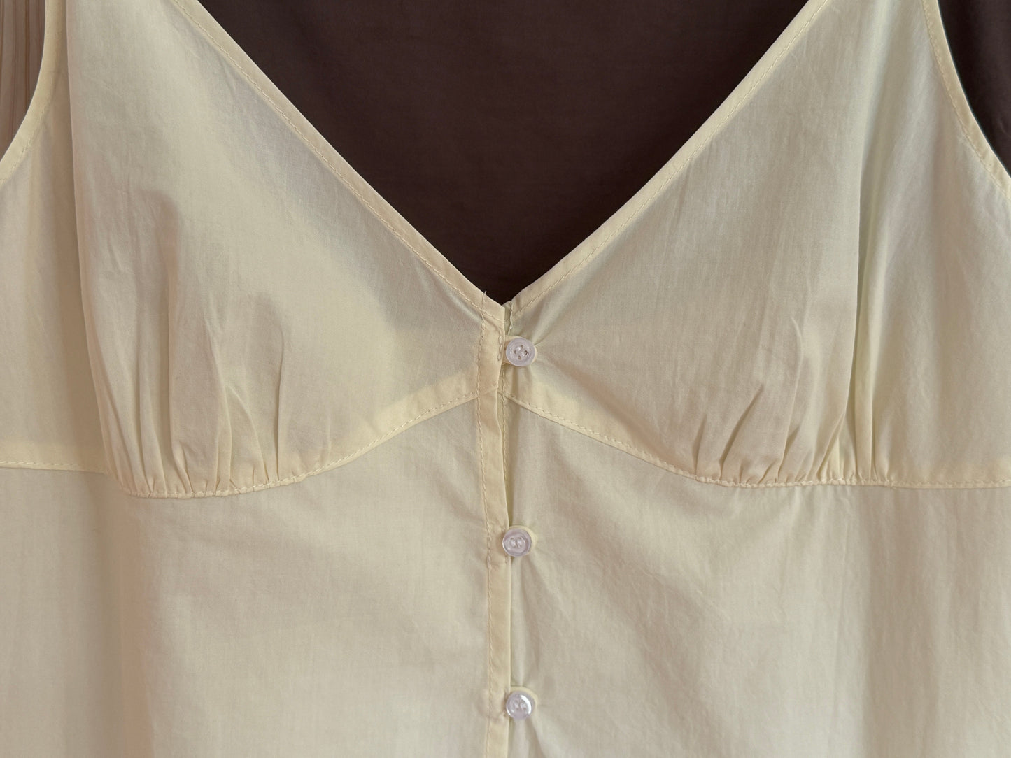 A close-up of the Double Seam Dress - Butter White by 0, a light yellow organic cotton sleeveless dress with a V-neckline and white front buttons, displayed against a dark background.