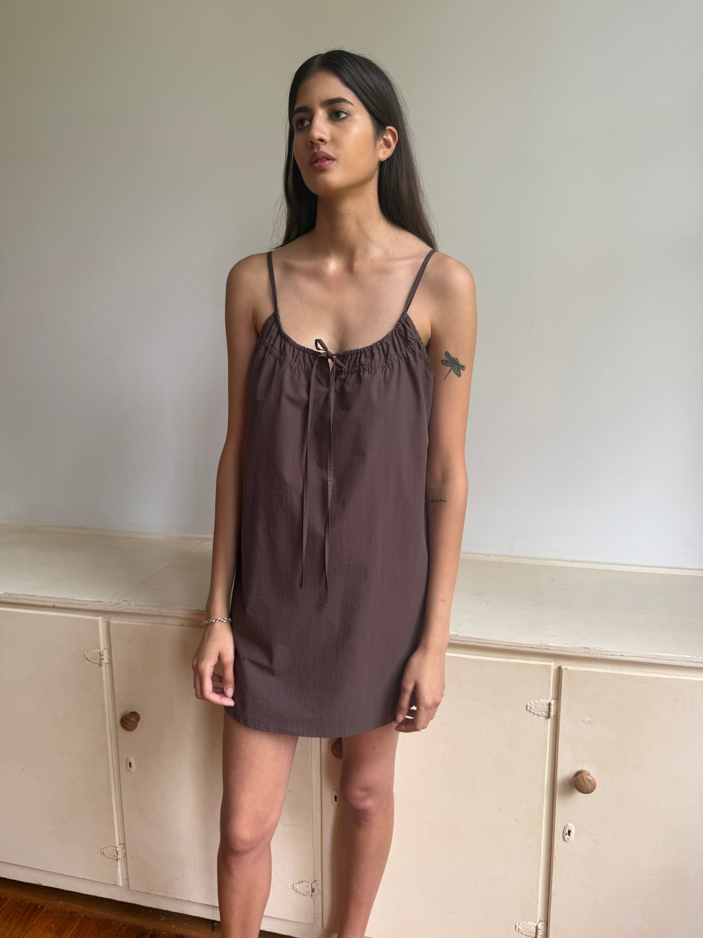 A woman with long dark hair stands indoors by a cream cabinet, wearing the Light Dress - Chocolate by 0—a sleeveless, mini brown dress with thin straps and a gathered neckline—and showing a small bird tattoo on her upper arm.