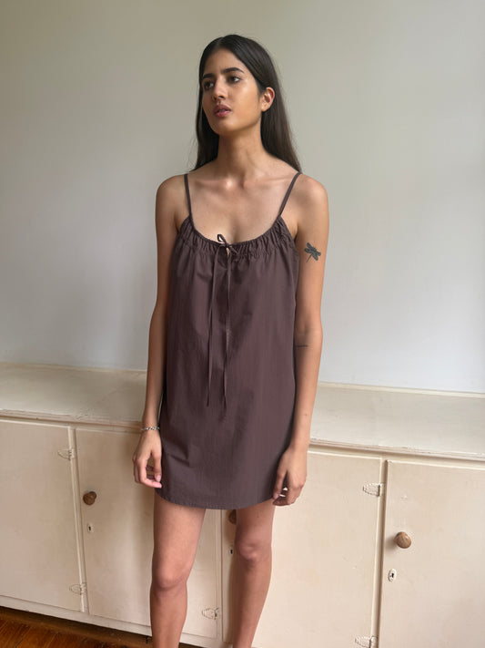 A woman with long dark hair stands indoors by a cream cabinet, wearing the Light Dress - Chocolate by 0—a sleeveless, mini brown dress with thin straps and a gathered neckline—and showing a small bird tattoo on her upper arm.