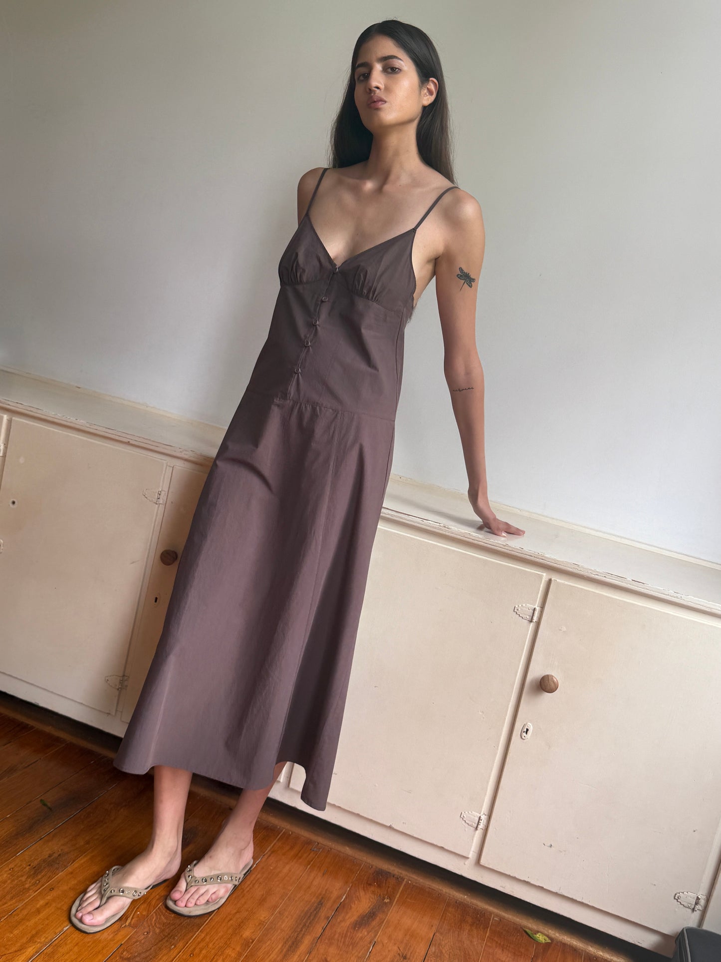 A woman with long dark hair, sandals, and an upper left arm tattoo stands indoors on a wooden floor, leaning against a cream cabinet. She wears the Double Seam Dress - Chocolate by 0, which features thin straps.