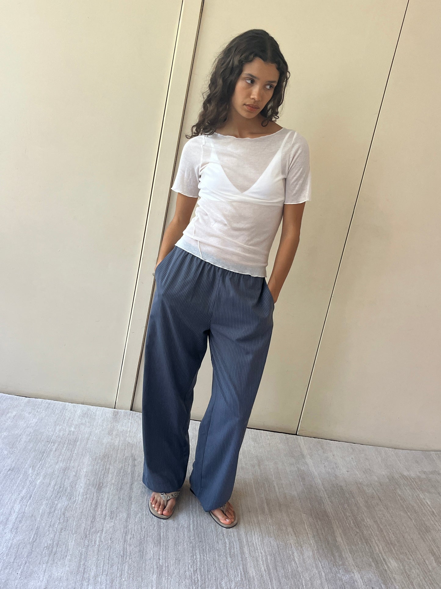 A woman stands against a beige wall wearing the Pure Wool Ease Trouser, paired with a sheer white short-sleeve top and sandals. Her hands are in her pockets as she gazes slightly aside with a neutral expression.
