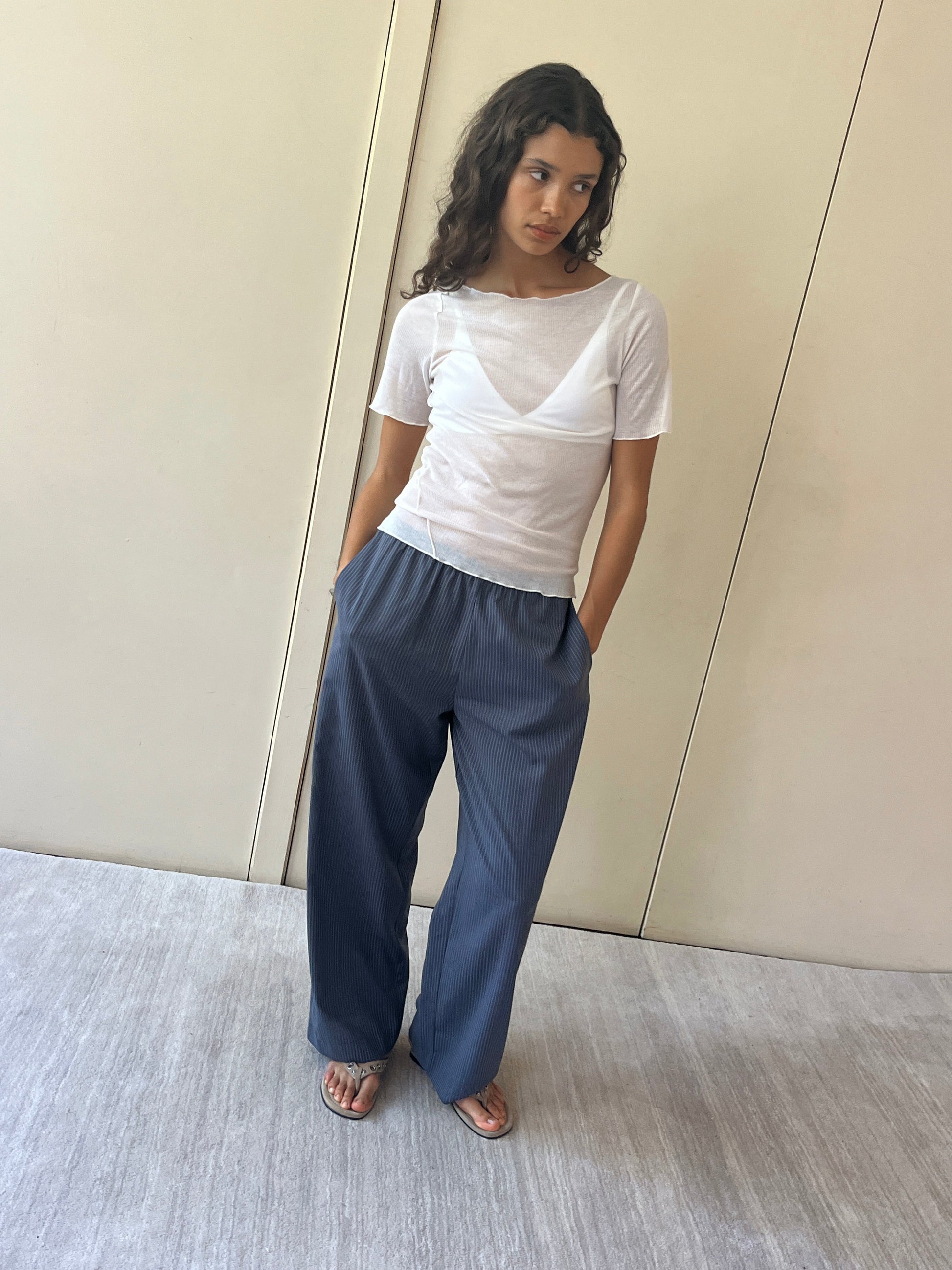 A woman stands against a beige wall wearing the Pure Wool Ease Trouser, paired with a sheer white short-sleeve top and sandals. Her hands are in her pockets as she gazes slightly aside with a neutral expression.