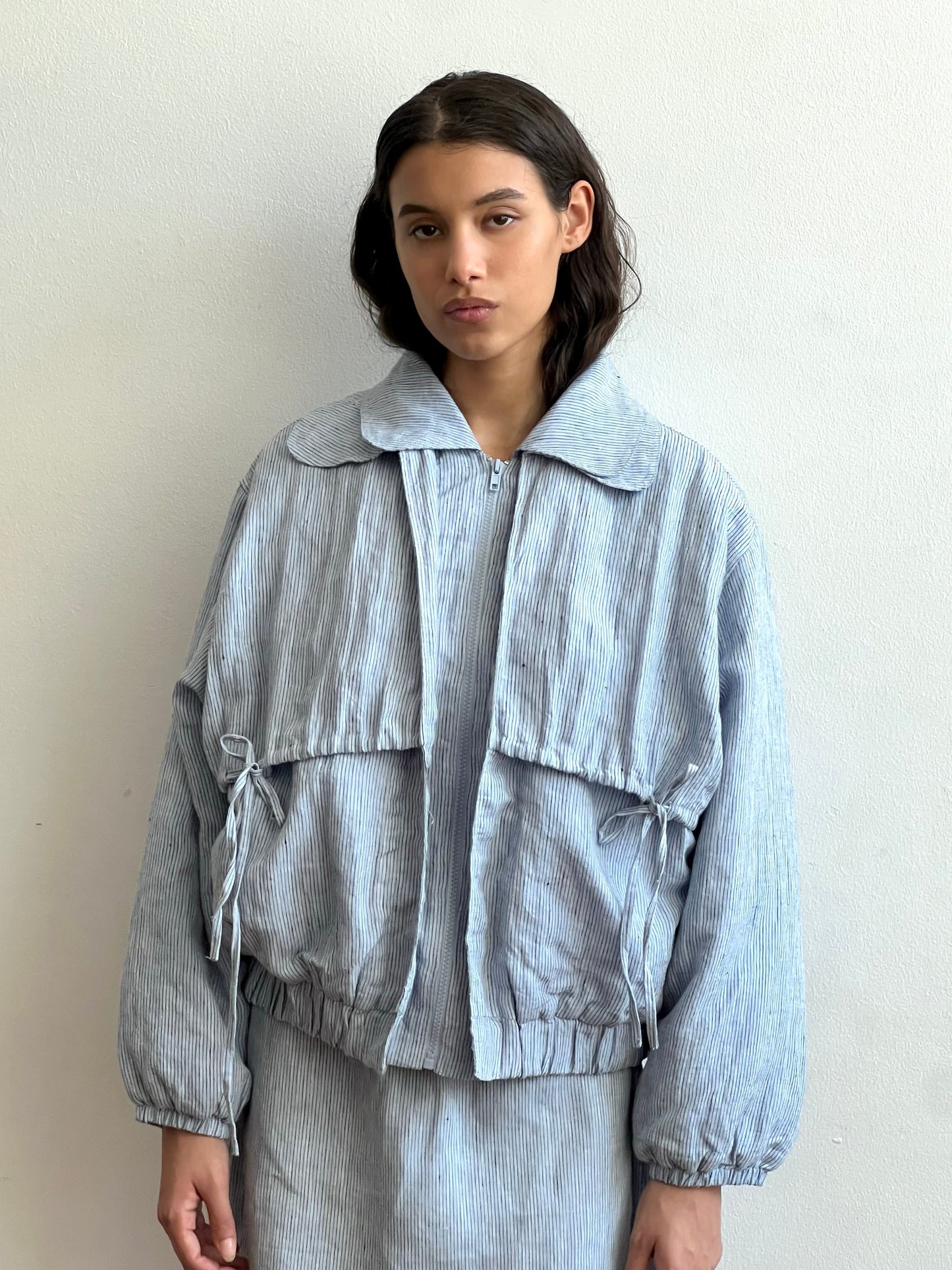 A woman with long dark hair stands against a white wall, wearing the Double Layered Jacket - Blue Stripe, a loose-fitting piece with large front pockets and matching pants. She keeps a neutral expression with her hands at her sides.