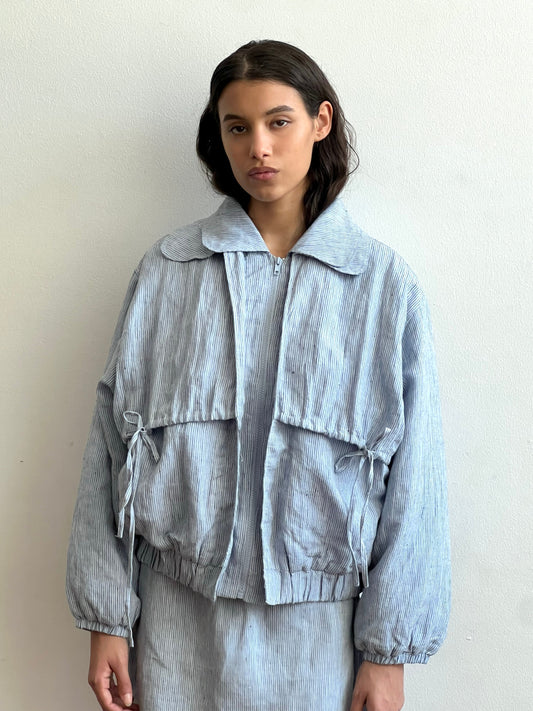 A woman with long dark hair stands against a white wall, wearing the Double Layered Jacket - Blue Stripe, a loose-fitting piece with large front pockets and matching pants. She keeps a neutral expression with her hands at her sides.