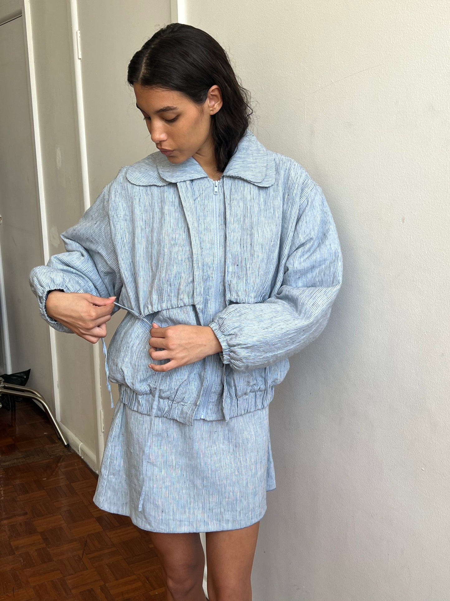 A woman in a light blue textured jacket and Deiji Studios Babylock Skirt - Blue Stripe stands indoors by a white wall and wooden floor, adjusting her jacket’s drawstring while looking down.