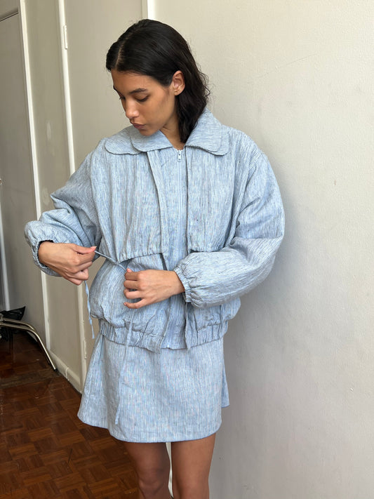 A woman in a light blue textured jacket and Deiji Studios Babylock Skirt - Blue Stripe stands indoors by a white wall and wooden floor, adjusting her jacket’s drawstring while looking down.