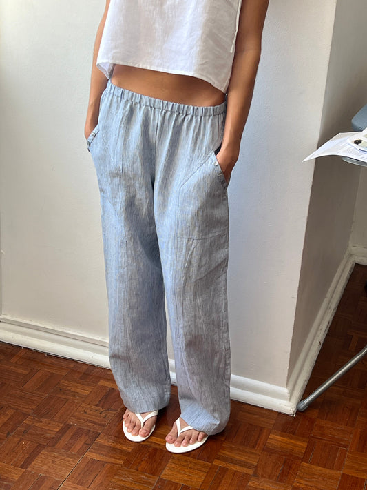 A person stands indoors on a wooden floor wearing the Angle Pocket Pant in light blue stripes, a white sleeveless crop top, and white flip-flops, with their hands in their pockets.