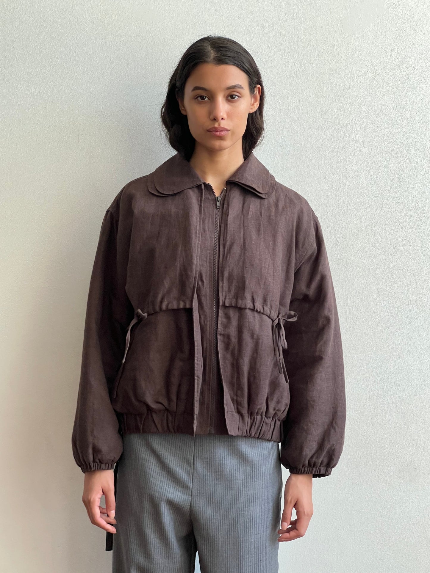 A person with long dark hair and a neutral expression stands against a light wall, wearing the Deiji Studios Double Layered Jacket - Bark with a zipper and elastic cuffs, paired with gray pants.