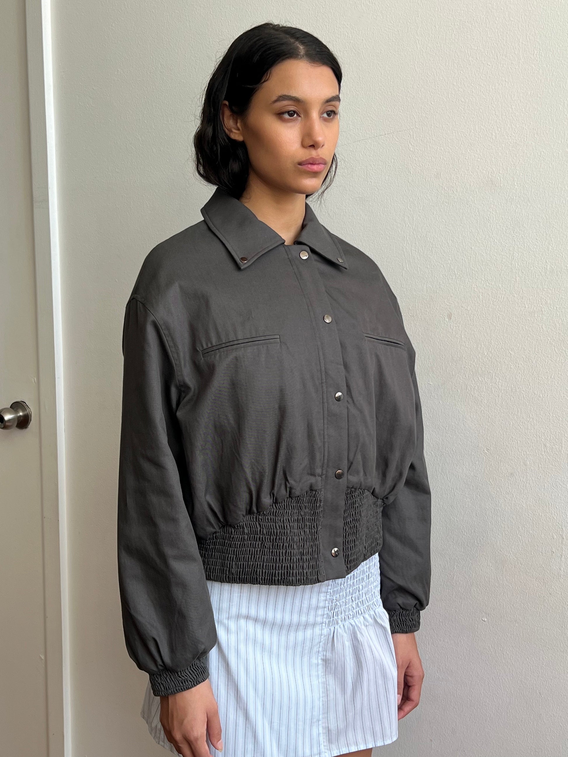 A person with dark hair wears the Deiji Studios Shirring Jacket - Slate with a smocked hem and cuffs, paired with a white and blue pinstriped skirt, standing indoors against a plain light-colored wall.