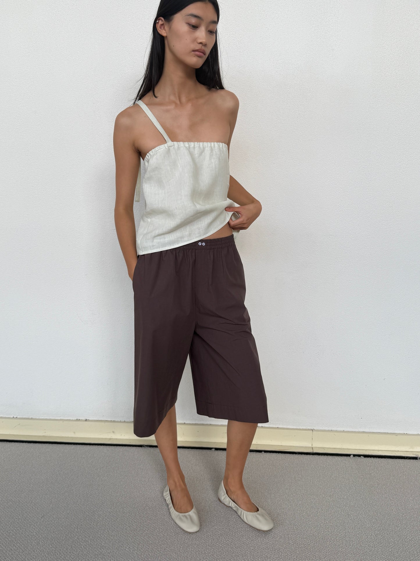 A woman stands against a white wall, wearing the Long Lounger - Chocolate shorts by 0, styled with a light beige asymmetrical strap top and beige flats. She looks to the side with her hands in her pockets.