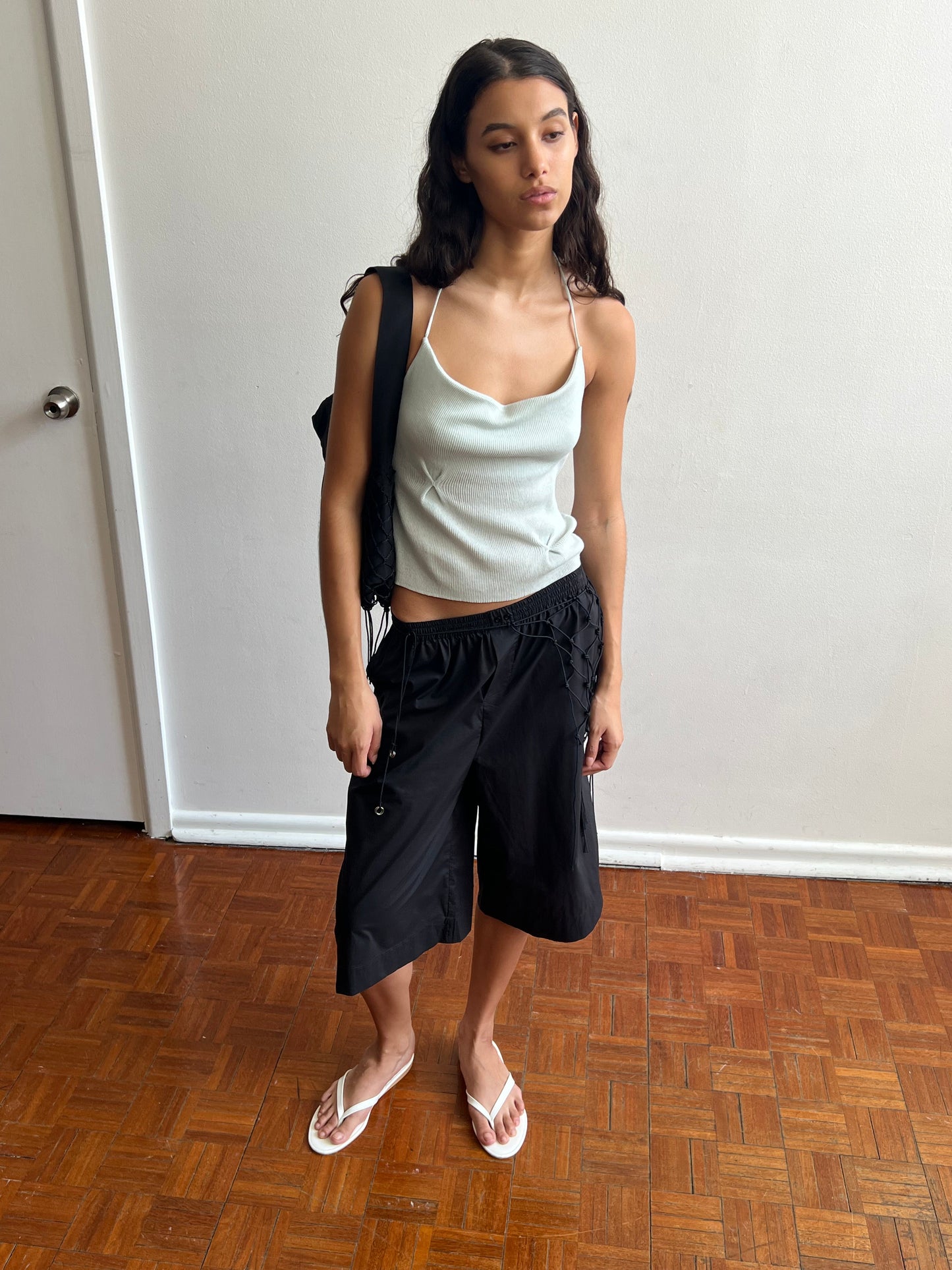 A woman with long dark hair and a neutral expression stands indoors on a wooden floor, wearing the Long Lounger, black knee-length shorts, white flip-flops, and a black backpack.