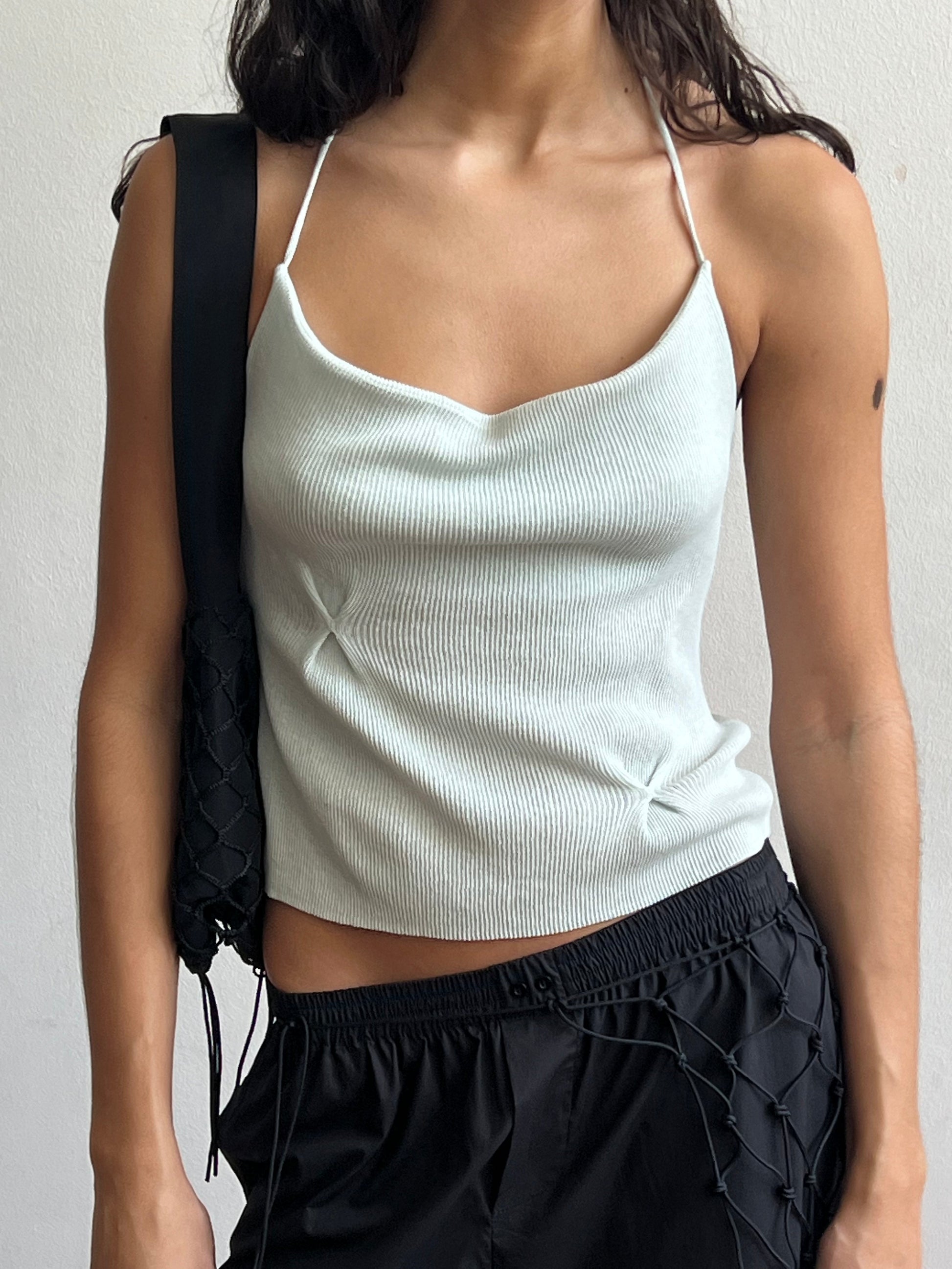 A woman wearing a Fine Knot Tank in ice blue and black drawstring pants with a black woven shoulder bag stands against a plain white background.