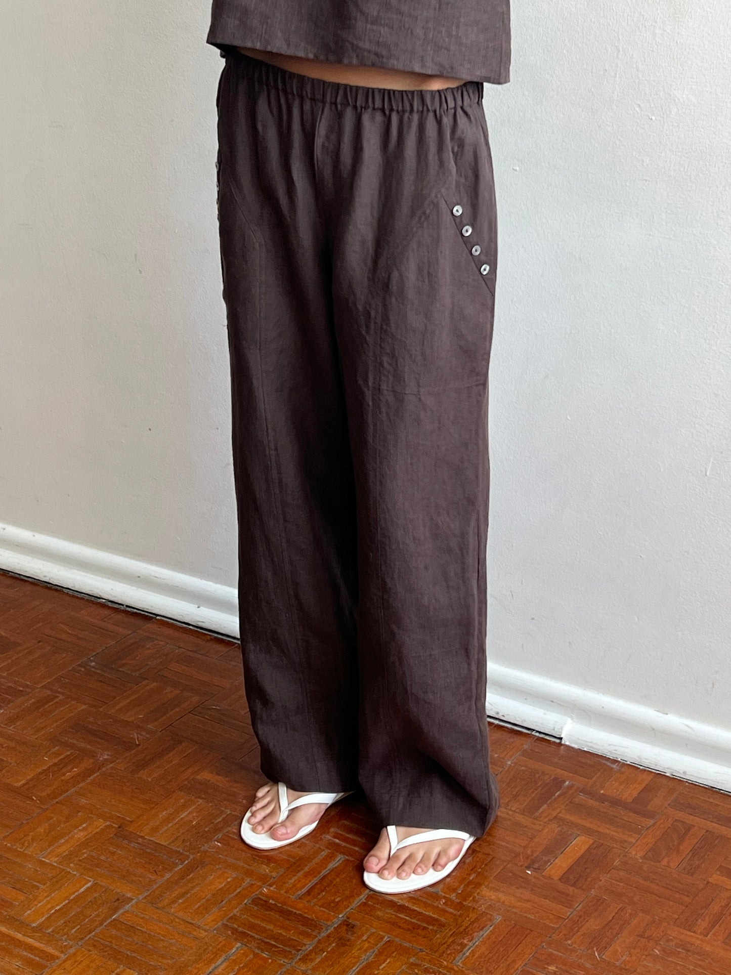 A person wearing the Angle Pocket Pant - Bark from Deiji Studios, featuring side button details, and white sandals stands on a wooden floor against a light-colored wall. Only the lower half of their body is visible.