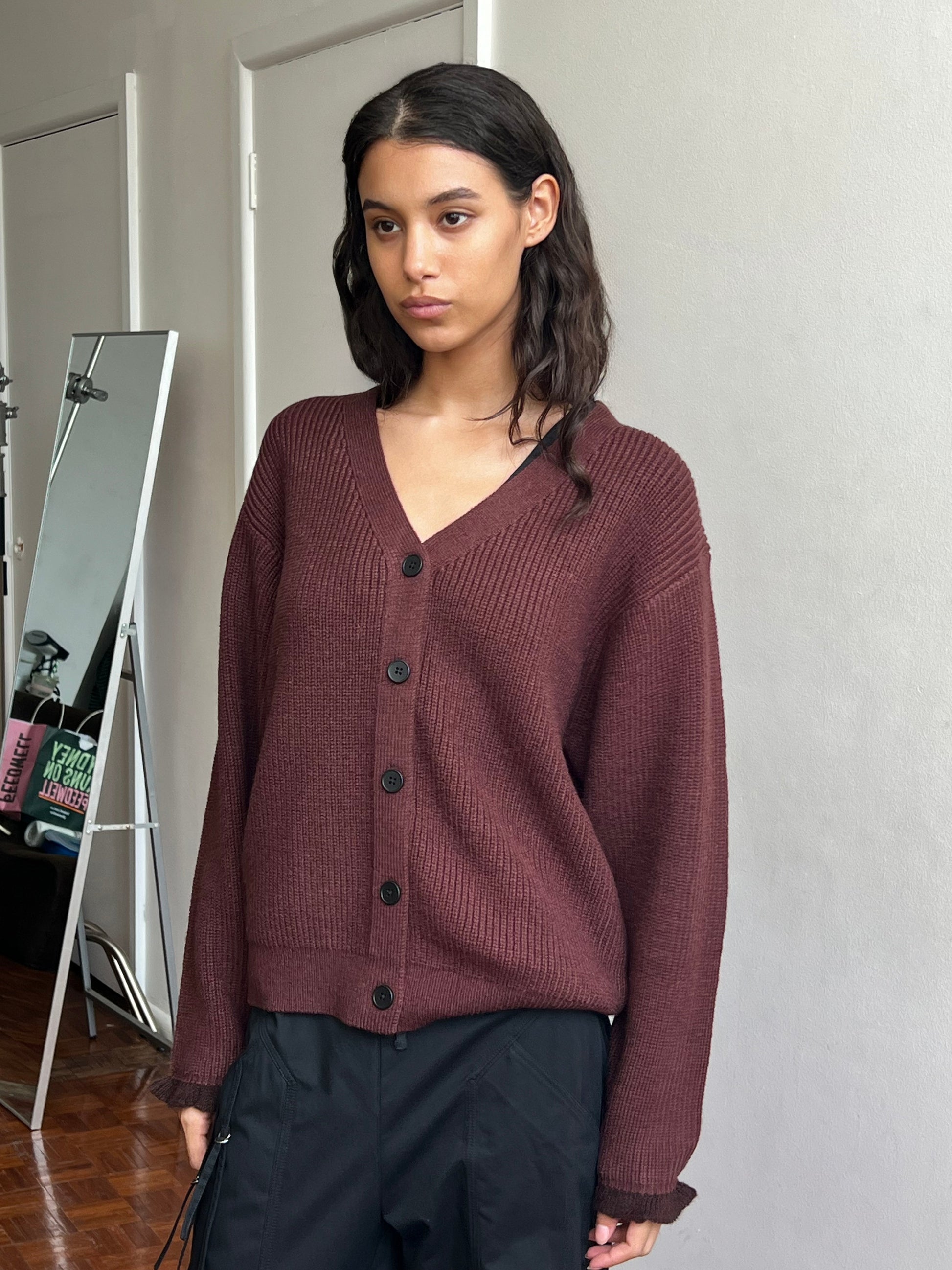 A woman with long dark hair stands indoors near a wall and a large mirror, wearing the Flounce Cardigan - Burgundy and black pants.