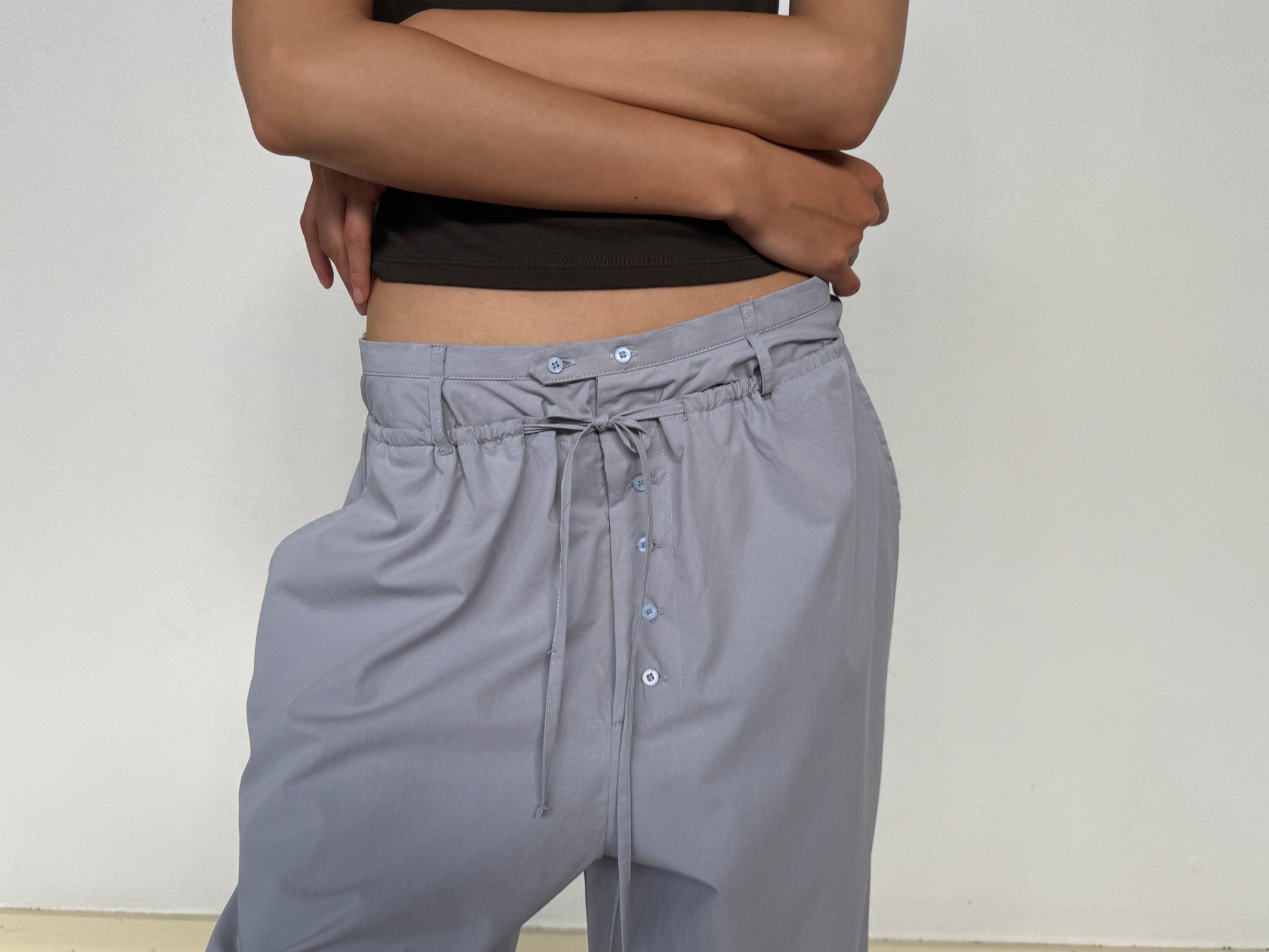 A person stands with arms crossed, wearing a dark sleeveless top and the Two Band Pant - Cornflower by 0, featuring light gray fabric, mid rise, drawstring waist, and visible buttons, set against a plain light background.