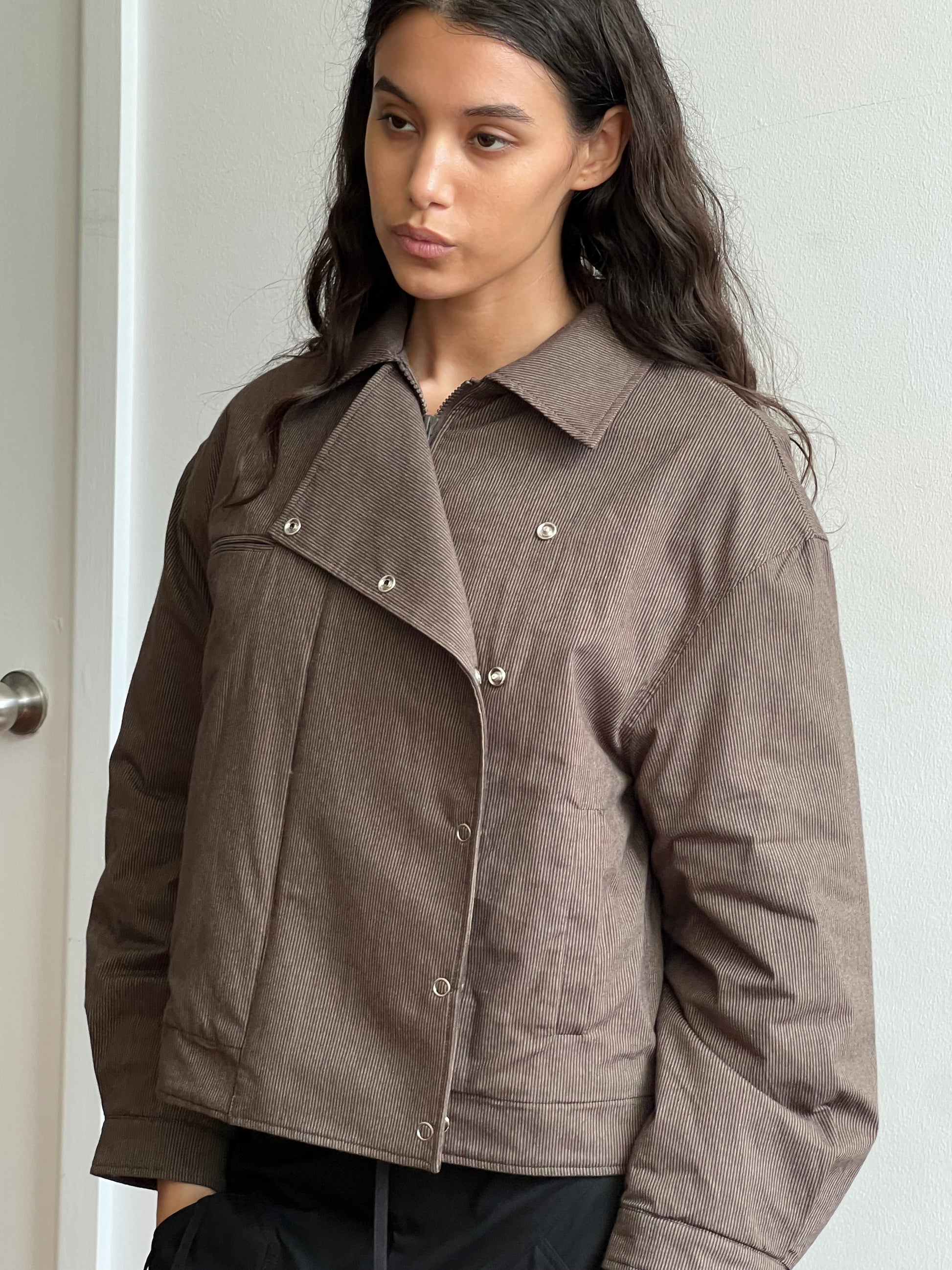 A woman with long brown hair wears the Lapel Jacket—a brown corduroy piece with snap buttons—standing indoors against a light wall and gazing slightly to the side with a neutral expression.