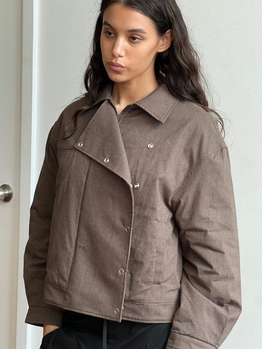A woman with long brown hair wears the Lapel Jacket—a brown corduroy piece with snap buttons—standing indoors against a light wall and gazing slightly to the side with a neutral expression.
