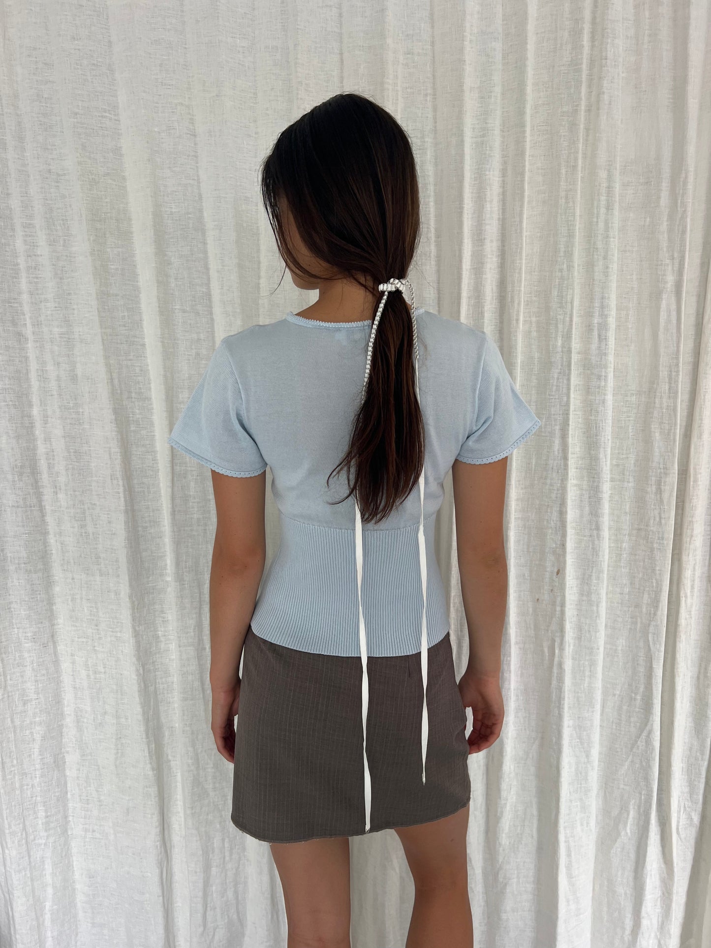 A woman faces away, wearing the Trim Knit Tee - Ice Blue from Deiji Studios and a brown skirt. Her long hair is tied back with a white beaded accessory and two white ribbons, standing before a white curtain.