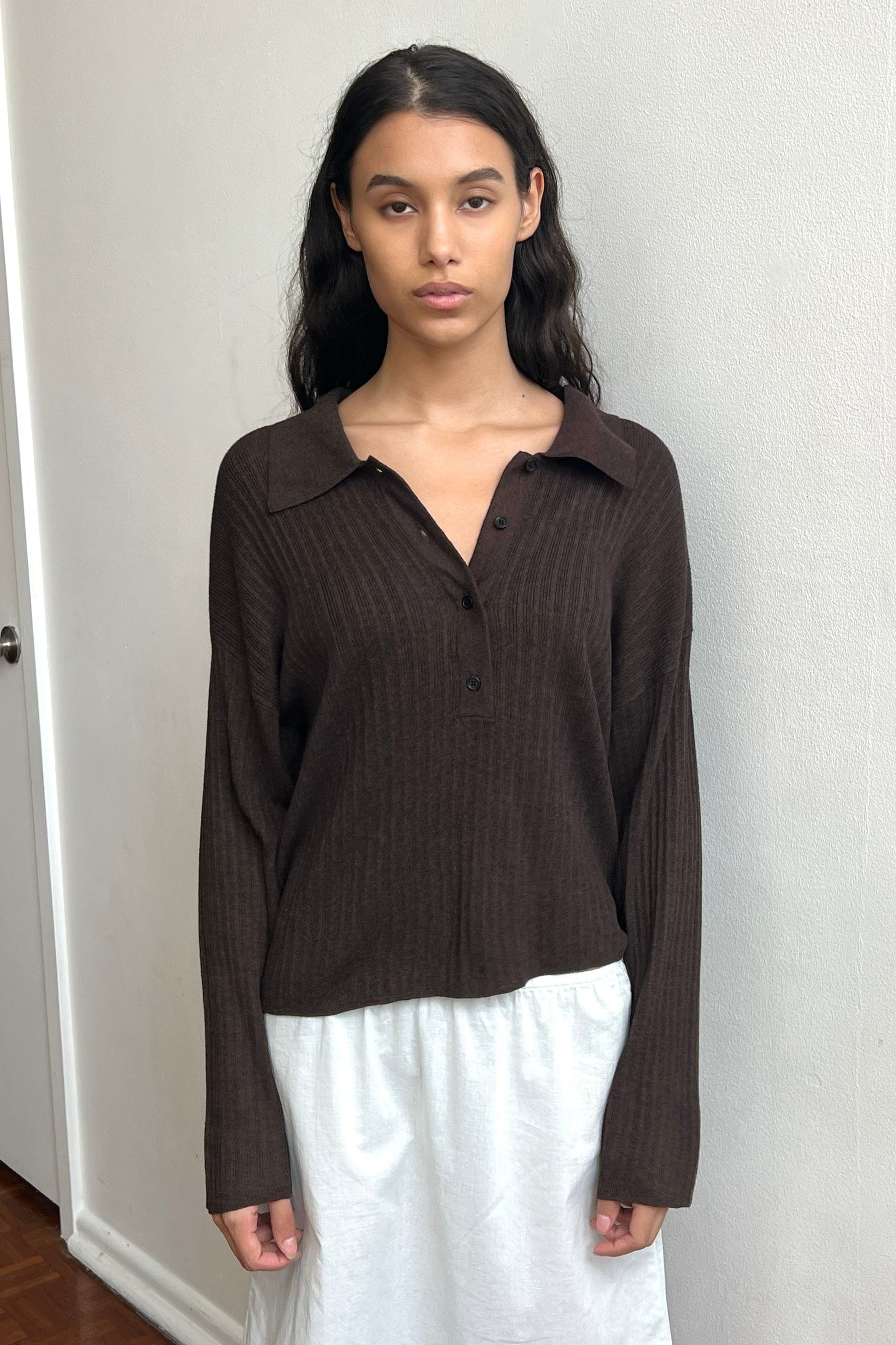 A young woman with long dark hair wears the Polo Knit, a loose, dark brown ribbed button-up top, paired with a white skirt. She stands indoors against a plain white wall, gazing directly at the camera with a neutral expression.
