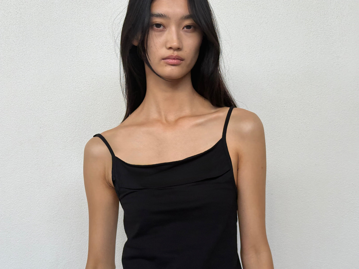 A person with long, straight black hair wears the 0 Simple Tank – Black, made from organic cotton, and stands against a plain white wall looking directly at the camera with a neutral expression.