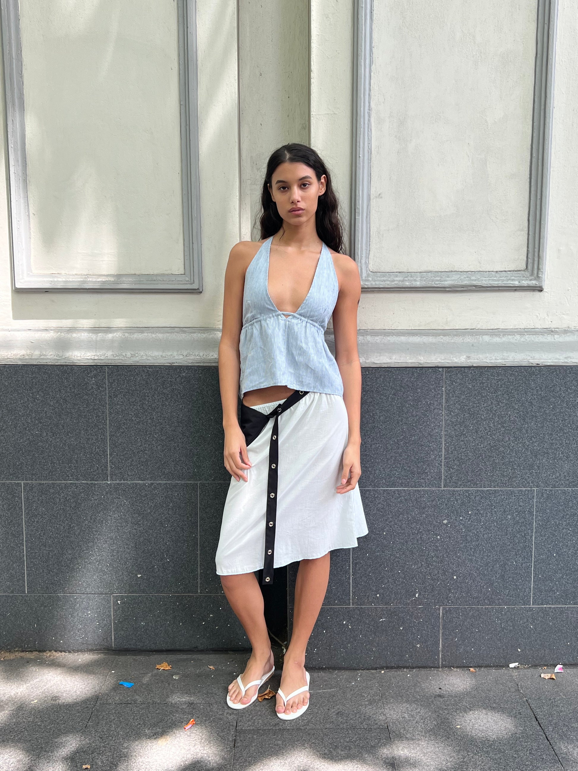 A woman with long dark hair stands against a gray and white wall, wearing the Linen Button Halter - Blue Stripe from Deiji Studios, a white wrap skirt with black buttons, and white flip-flops. She gazes neutrally at the camera.
