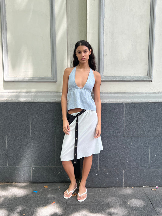 A woman with long dark hair stands against a gray and white wall, wearing the Linen Button Halter - Blue Stripe from Deiji Studios, a white wrap skirt with black buttons, and white flip-flops. She gazes neutrally at the camera.