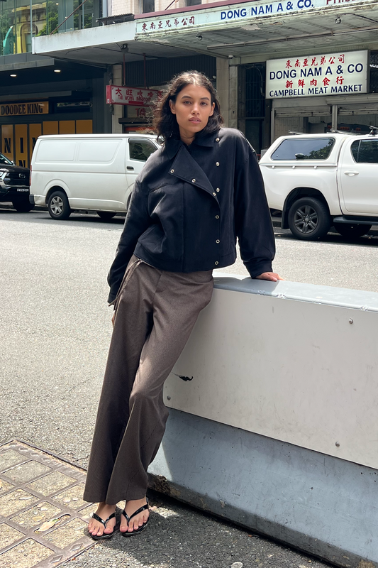 A woman leans against a street barrier wearing a black Lapel Jacket, brown wide-leg trousers, and black thong sandals. Behind her are city storefronts, including a meat market, and white vehicles.