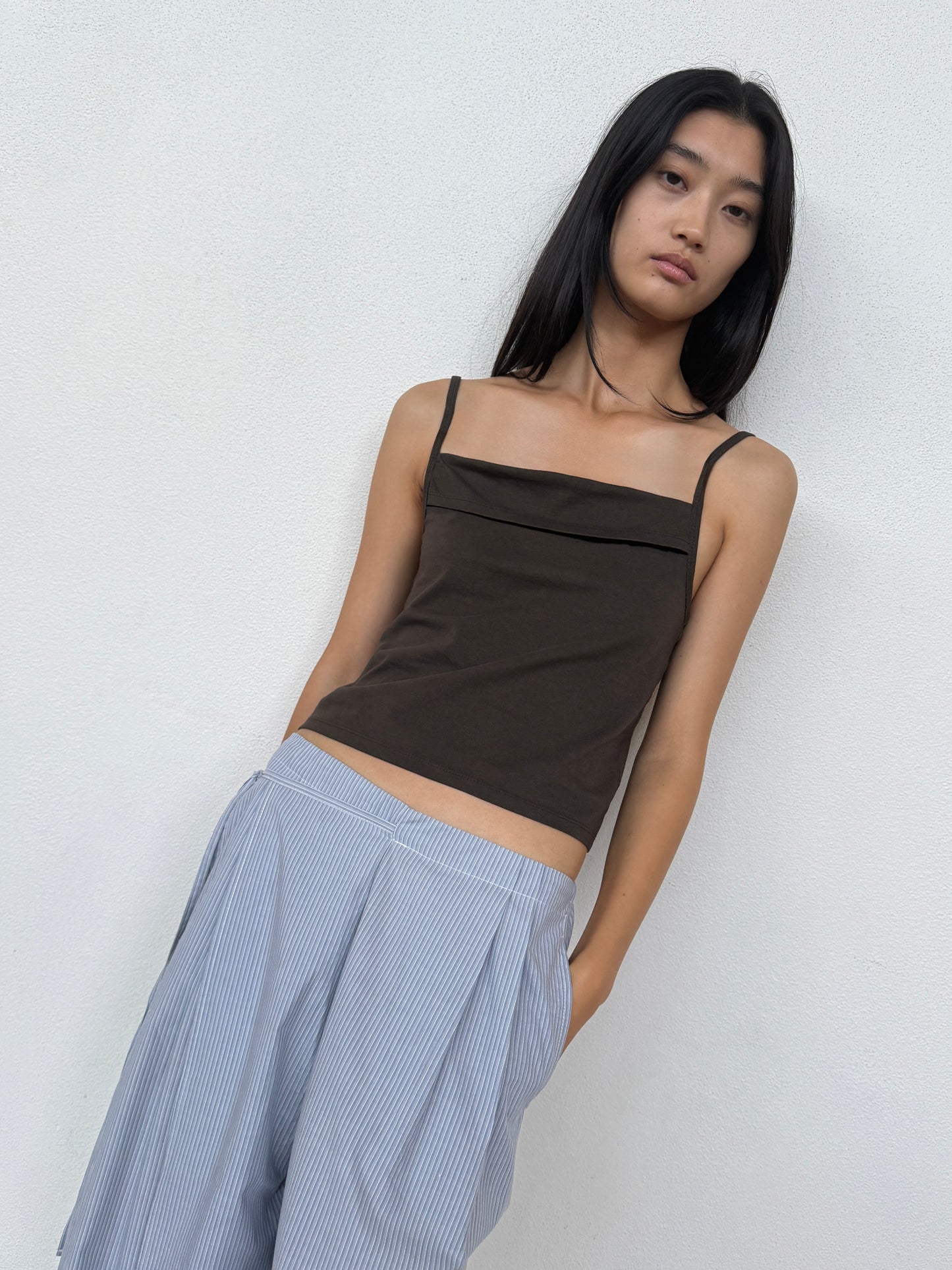 A person with long black hair wears the 0 Simple Tank - Caribou in dark brown and loose light blue striped pants, standing against a white wall and looking at the camera with a neutral expression.