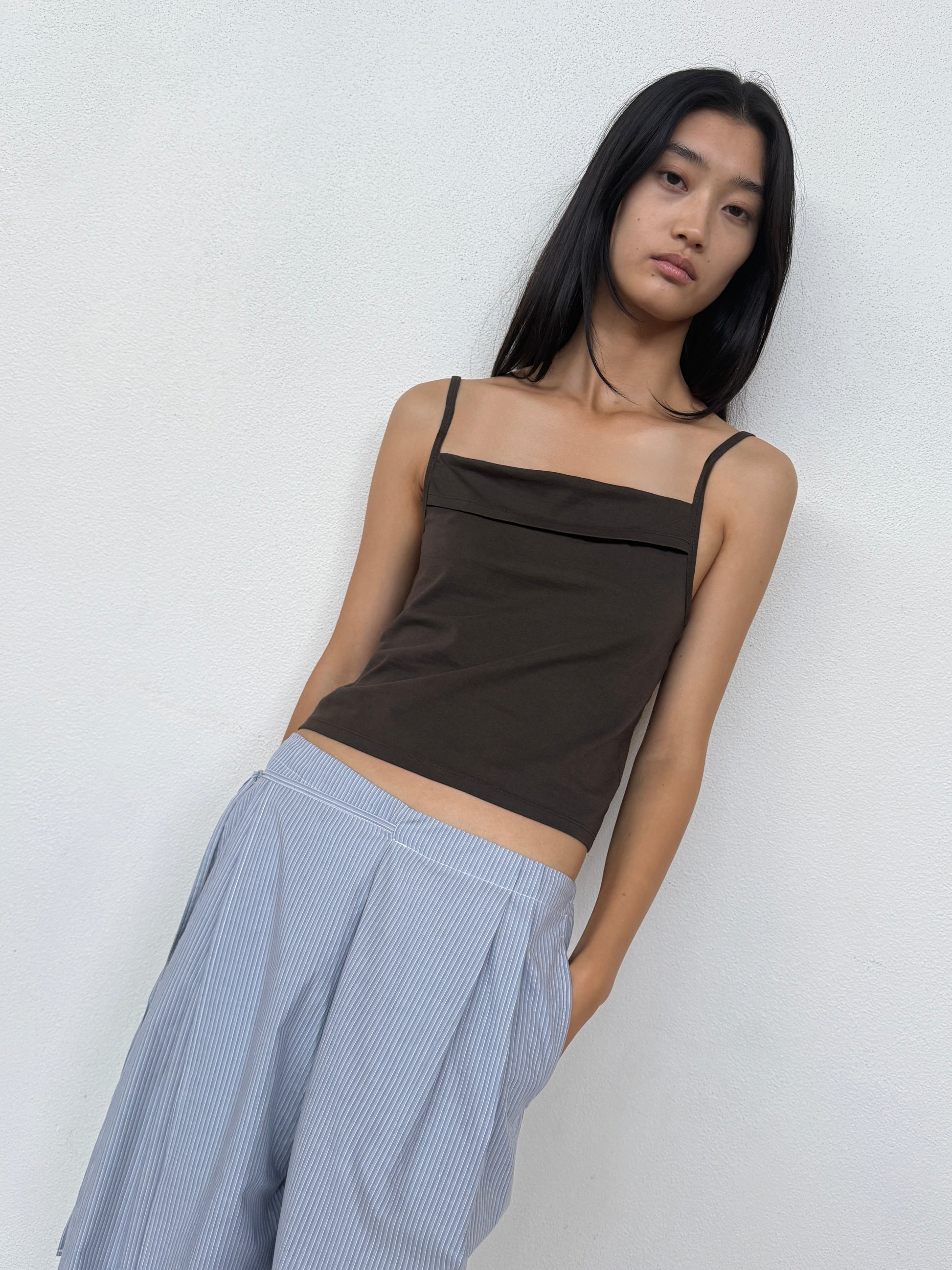 A person with long black hair wears the 0 Simple Tank - Caribou in dark brown and loose light blue striped pants, standing against a white wall and looking at the camera with a neutral expression.