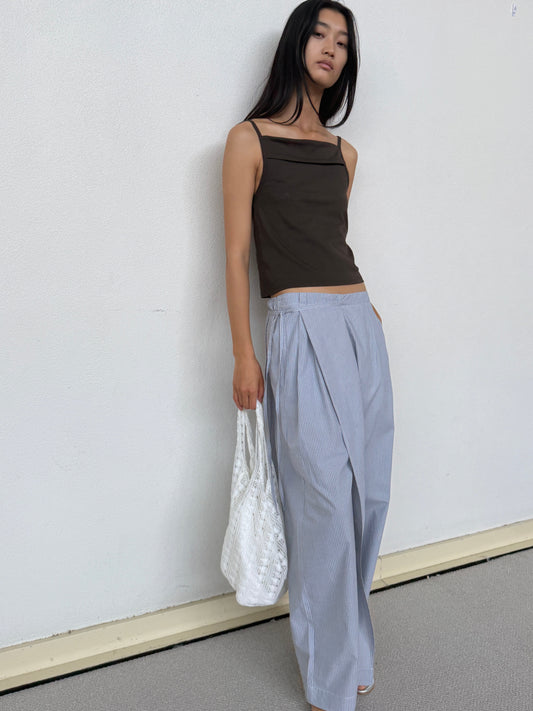 A person with long black hair stands against a white wall wearing a sleeveless dark top and loose, light blue pinstriped pants, holding the Thread Dust Bag - Off White on their shoulder and posing with a relaxed expression.