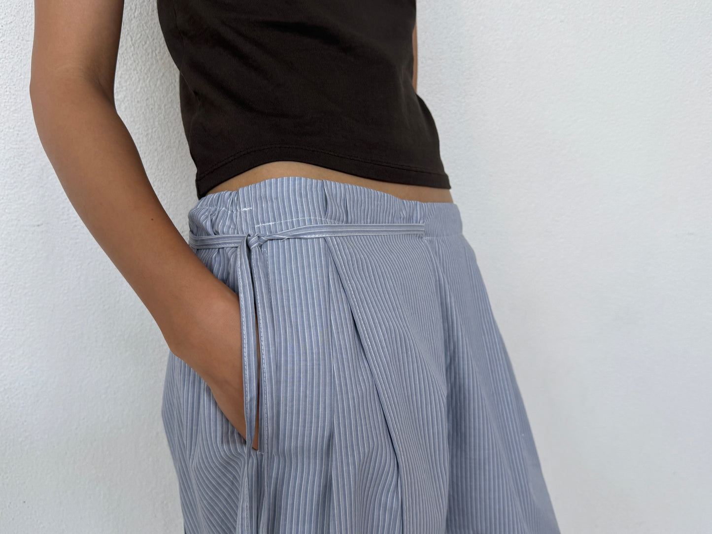 A person models the 0 Double Layer Pant - Sky Stripe and a black top against a white wall, with one hand in their pocket and the other relaxed at their side.