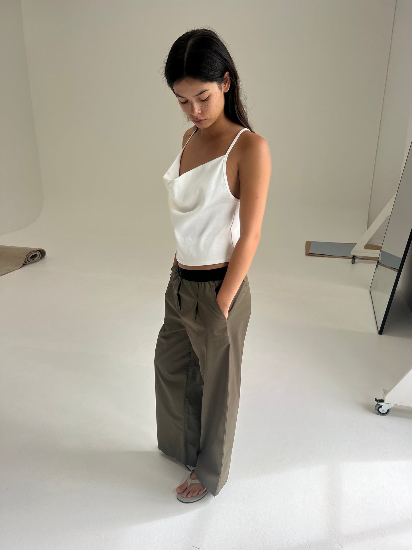 A woman with long dark hair stands indoors on a white floor, looking down. She wears a sleeveless top and the Deiji Studios Contrast Waist Pant - Olive. The minimal background includes neutral tones and some visible studio equipment.