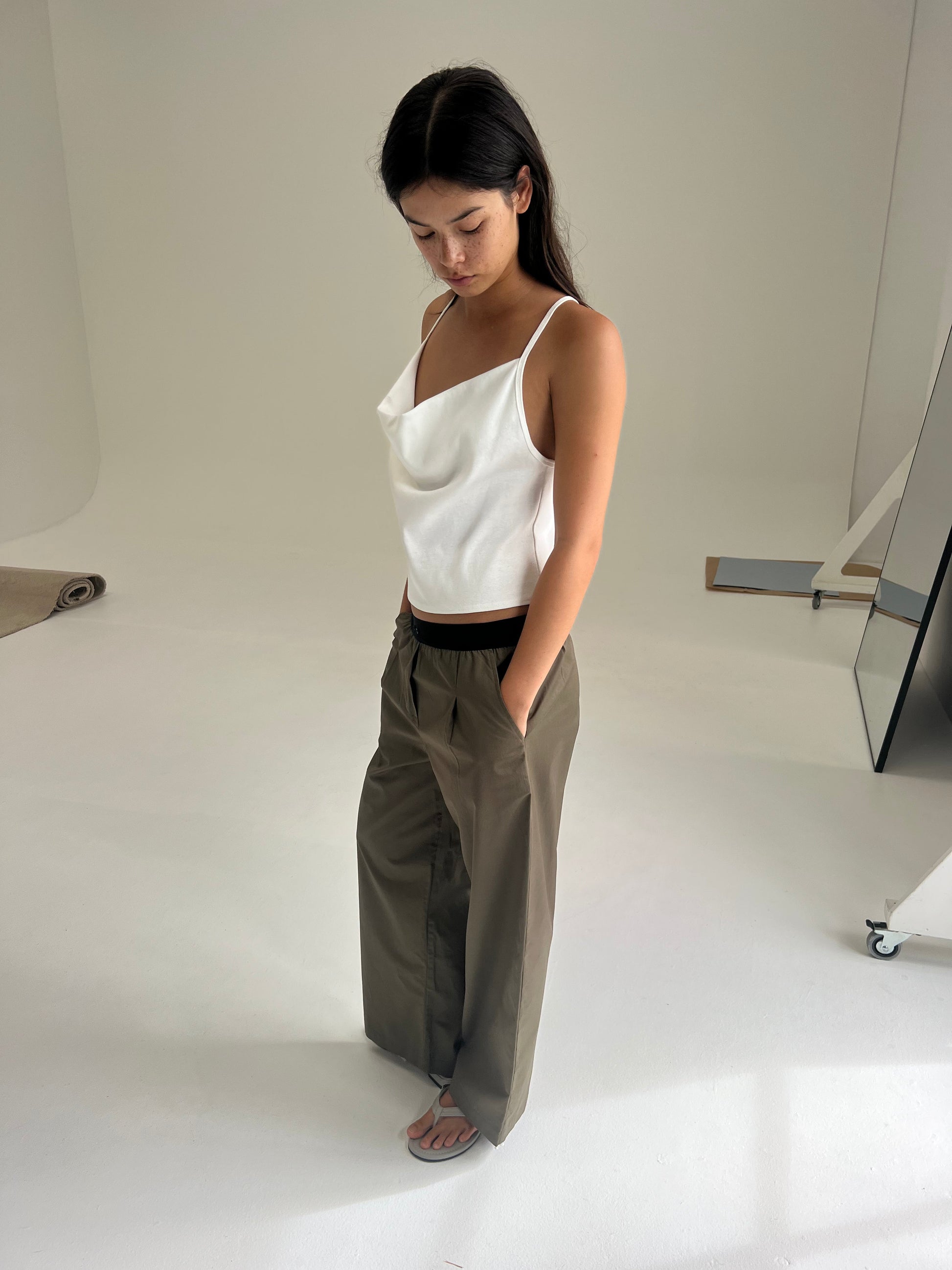 A woman with long dark hair stands indoors on a white floor, looking down. She wears a sleeveless top and the Deiji Studios Contrast Waist Pant - Olive. The minimal background includes neutral tones and some visible studio equipment.