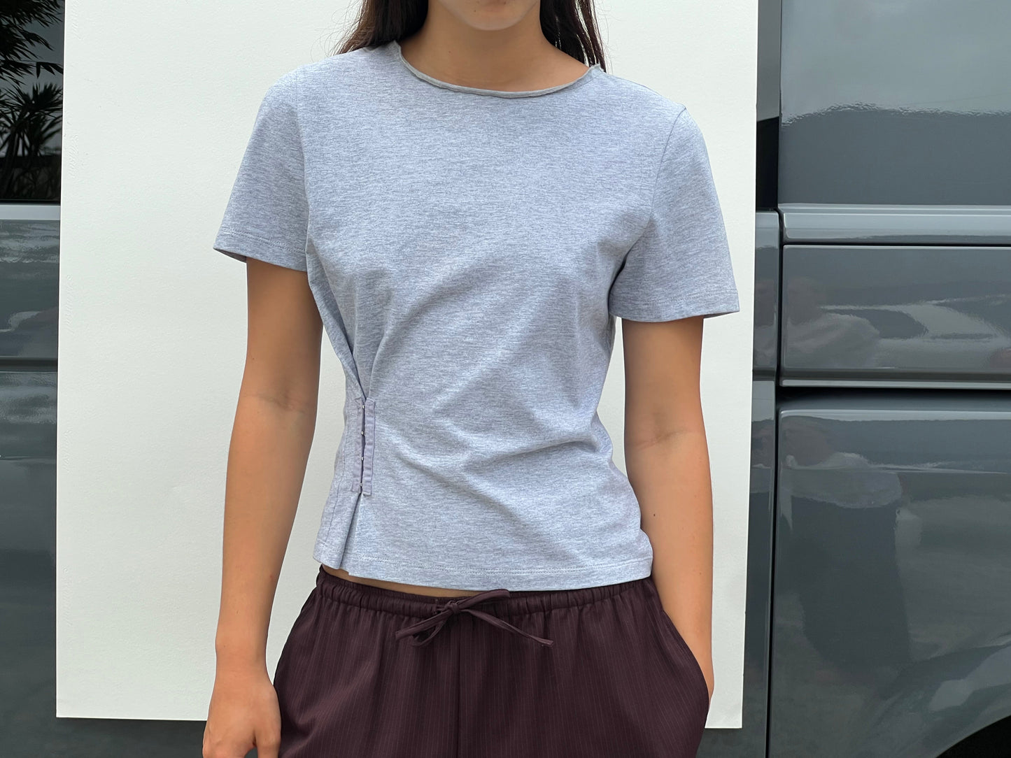 A woman stands outdoors wearing the Pinch Tee - Grey by Deiji Studios with dark brown drawstring pants; her head is partially out of frame, with a reflective surface and white panel in the background.
